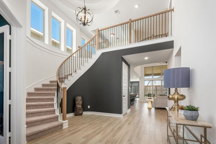 This photo showcases a modern entryway with a dramatic curved staircase and large windows providing ample natural light. The space features light wood flooring, a stylish chandelier, and an open view into the living area, creating an inviting and spacious atmosphere.