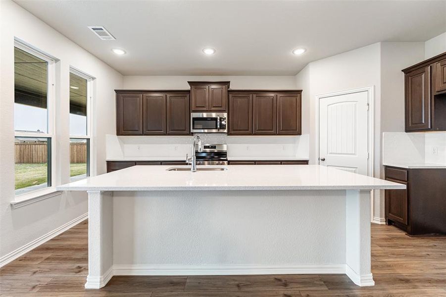 Kitchen with dark brown cabinets, light stone counters, a kitchen island with sink, light wood-style flooring, and recessed lighting
