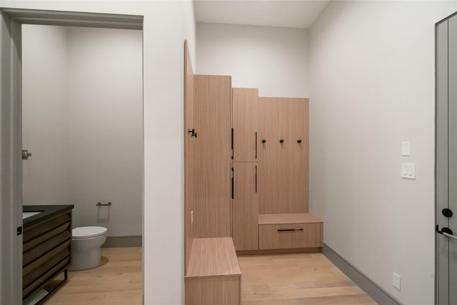 This photo shows a modern mudroom with light wood lockers and hooks for storage. Adjacent is a small bathroom with a toilet and a sleek vanity, featuring light wood flooring throughout.