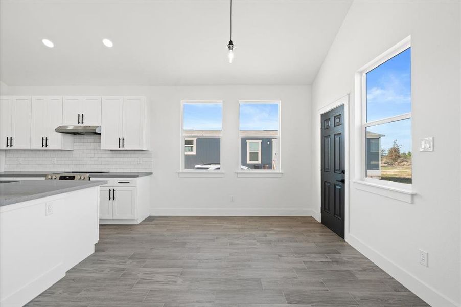 This is a bright, modern kitchen with white cabinetry, stainless steel appliances, subway tile backsplash, and gray flooring. The space is well-lit with natural light from multiple windows and has a high ceiling.