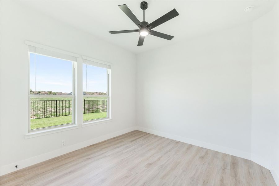Empty bedroom with light wood floors, black ceiling fan, and two windows overlooking a green yard