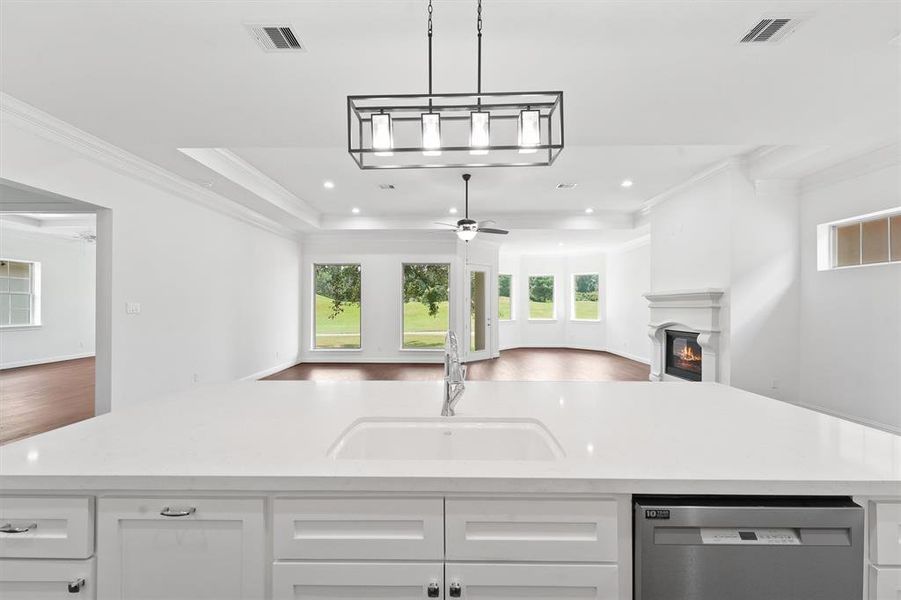 This kitchen features a large island overlooking an open-concept living area with a fireplace and views of a green outdoor space through multiple windows.