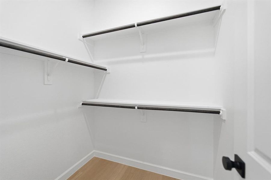 White walk-in closet featuring light wood-finish flooring, white walls, and multiple built-in shelving units with black hanging rods