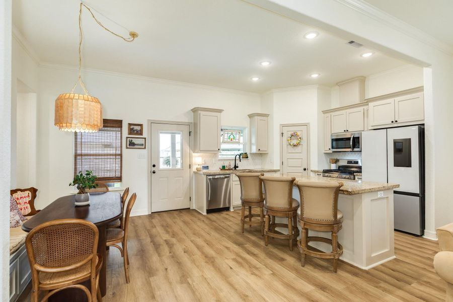 This bright kitchen features modern appliances, light cabinetry, and a spacious island with seating. The adjacent dining area offers cozy bench seating and a warm, inviting atmosphere with wood look flooring throughout.