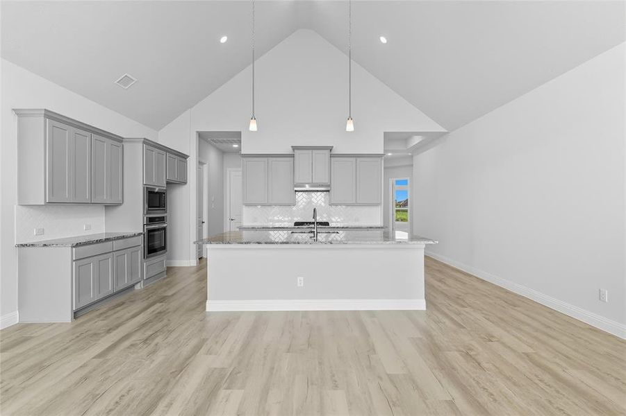 Kitchen with gray cabinets, high vaulted ceiling, light stone countertops, tasteful backsplash, and a kitchen island with sink Kitchen with gray cabinets, high vaulted ceiling, light stone countertops, tasteful backsplash, and a kitchen island with sink