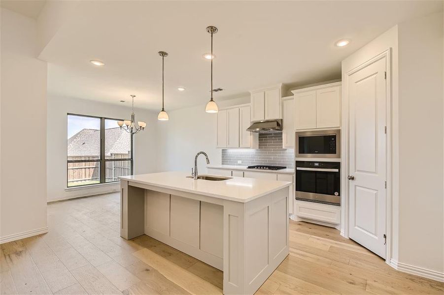 Kitchen with decorative light fixtures, wall chimney range hood, backsplash, a kitchen island with sink, and appliances with stainless steel finishes