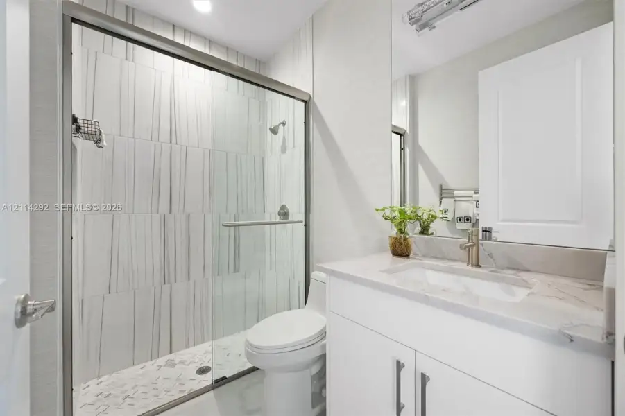 Bathroom featuring a walk-in glass shower with vertical-patterned wall tile, stone-surface countertop, integrated rectangular sink, vanity with white cabinetry, and brushed-metal hardware