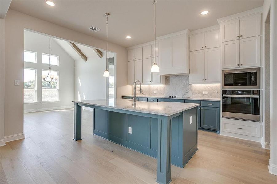 Kitchen featuring oven, white cabinets, an island with sink, light wood-type flooring, and vaulted ceiling with beams