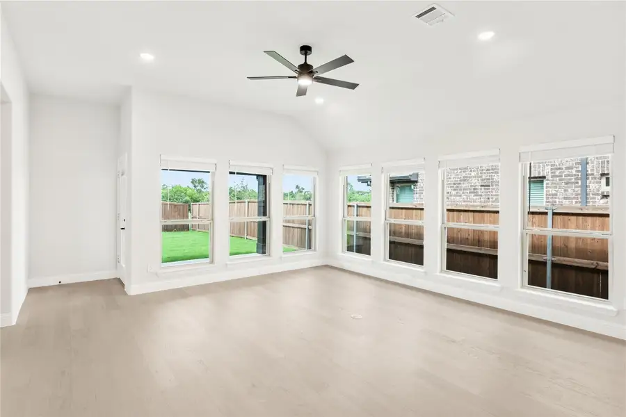 Empty living room with vaulted ceiling, ceiling fan, light wood floors, and large windows overlooking fenced backyard