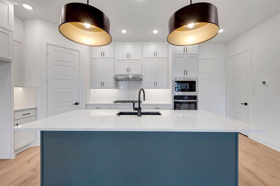 Kitchen featuring a large island with a white countertop and blue-finish base, dual contemporary pendant lighting, white shaker cabinetry, stainless steel appliances, and wood-finish flooring Kitchen featuring a large island with a white countertop and blue-finish base, dual contemporary pendant lighting, white shaker cabinetry, stainless steel appliances, and wood-finish flooring