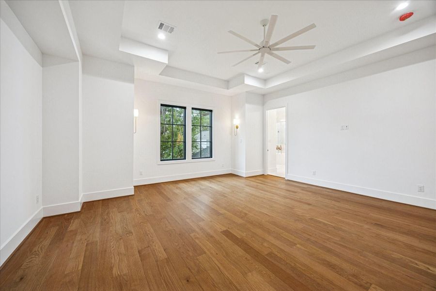 Tray-ceiling architecture, wide-plank oak floors, and soft natural light shape a serene and spacious primary suite. Wall sconces add understated warmth, while large black-framed windows capture tree-top views. The generous footprint easily accommodates multiple furniture layouts, offering flexibility for a private lounge or reading nook. Tray-ceiling architecture, wide-plank oak floors, and soft natural light shape a serene and spacious primary suite. Wall sconces add understated warmth, while large black-framed windows capture tree-top views. The generous footprint easily accommodates multiple furniture layouts, offering flexibility for a private lounge or reading nook.