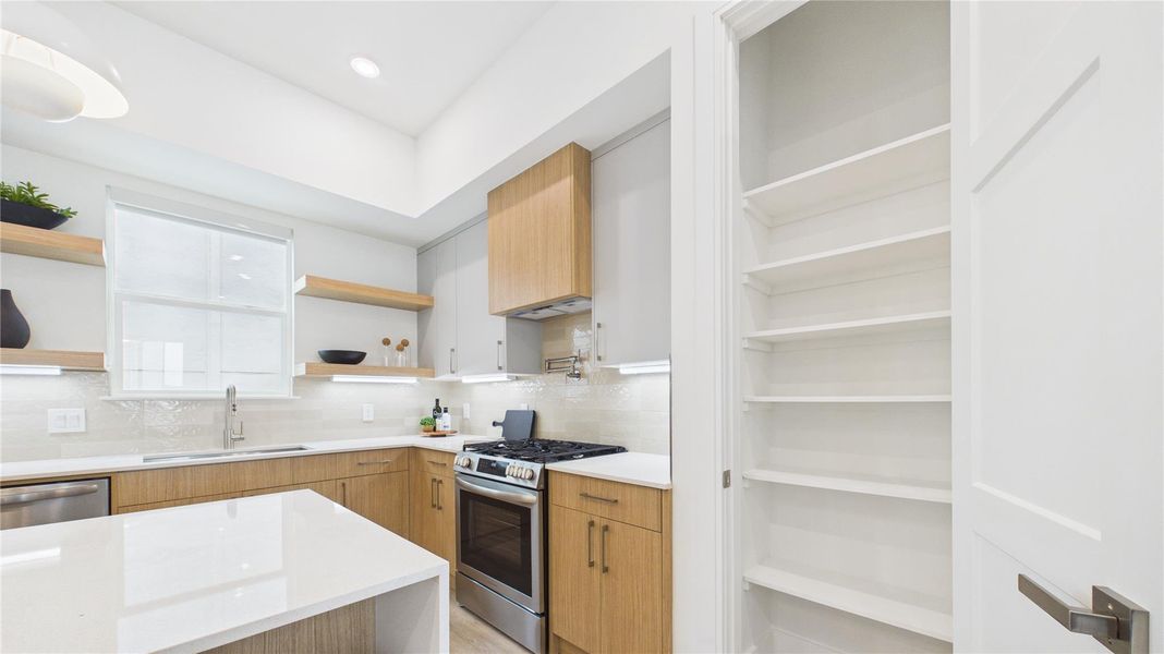 This modern kitchen features sleek wooden cabinetry, a spacious island, and stainless steel appliances. It includes open shelving, a window for natural light, and an adjacent pantry with ample shelving.