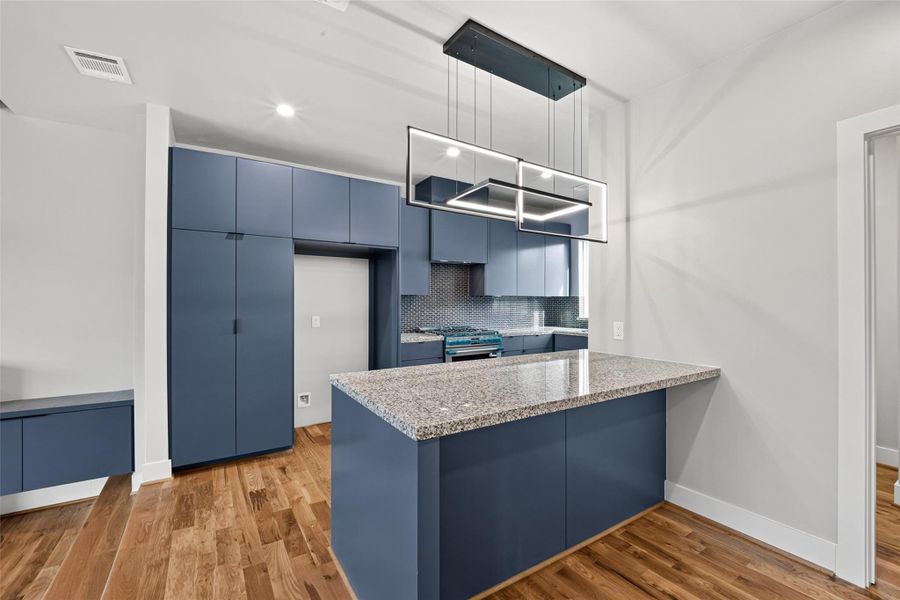 The chef's kitchen is a standout feature of this home. Bold blue flat-front cabinetry runs floor to ceiling, complemented by granite countertops, mosaic tile backsplash, stainless steel gas range, and a large peninsula island with seating . Hardwood floors and spectacular modern lighting complete the look.