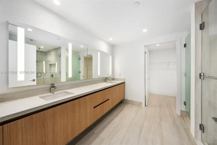 Floating wood-finish vanity with dual integrated sinks, expansive wall-to-wall mirror with integrated LED lighting, stone-finish flooring, and glass-enclosed shower Floating wood-finish vanity with dual integrated sinks, expansive wall-to-wall mirror with integrated LED lighting, stone-finish flooring, and glass-enclosed shower