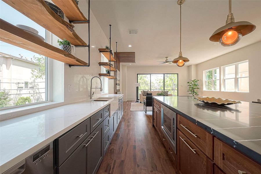 This modern kitchen features sleek dark cabinetry, white countertops, wooden open shelving, and pendant lighting, with a spacious layout that flows into a bright living area. This modern kitchen features sleek dark cabinetry, white countertops, wooden open shelving, and pendant lighting, with a spacious layout that flows into a bright living area.