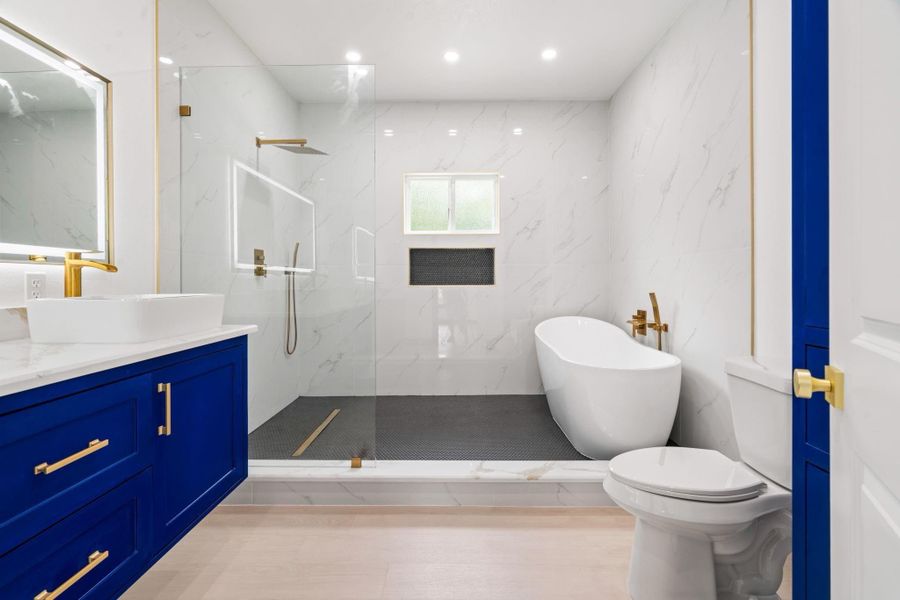 A luxurious primary bathroom showcases a spacious glass-enclosed shower, a sleek freestanding tub, and a sophisticated blue floating vanity. Gold fixtures, marble wall finishes, and recessed lighting complete the upscale, modern ambiance.