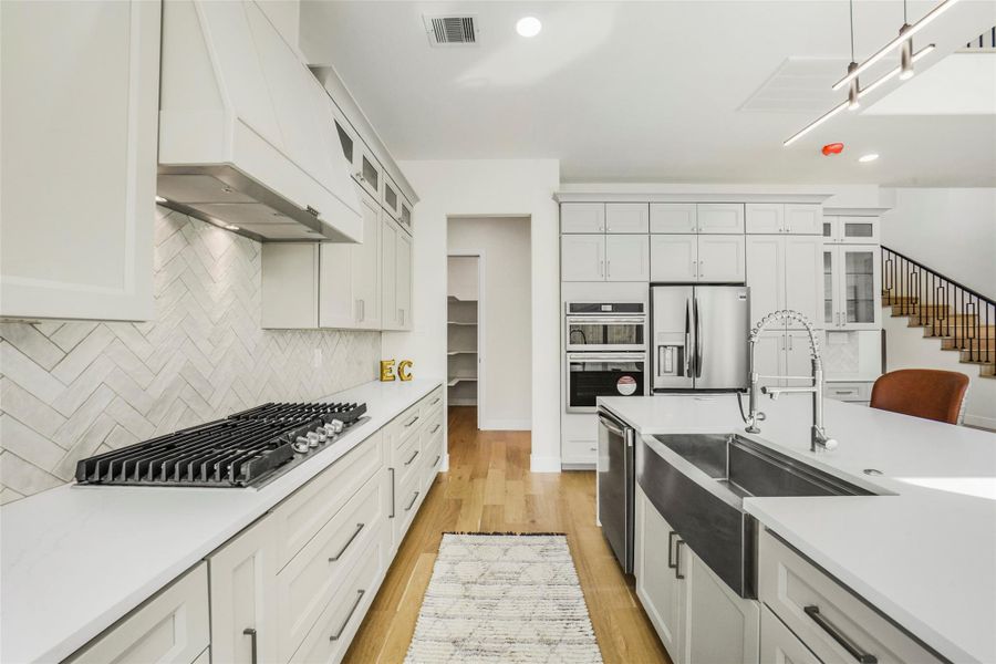 This modern kitchen features sleek white cabinetry, a stainless steel double oven and fridge, a spacious island with a farmhouse sink, gas cooktop, and stylish herringbone backsplash. It's open and bright, perfect for cooking and entertaining.