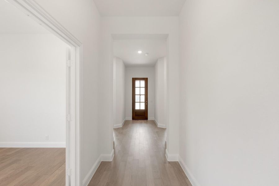 Hallway with white walls, light wood floors, and wood front door with glass panels at the end