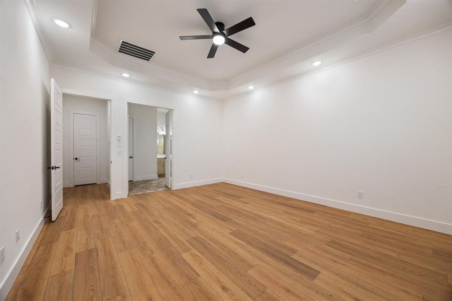 Unfurnished bedroom featuring a raised ceiling, ensuite bath, light wood-type flooring, ornamental molding, and a ceiling fan