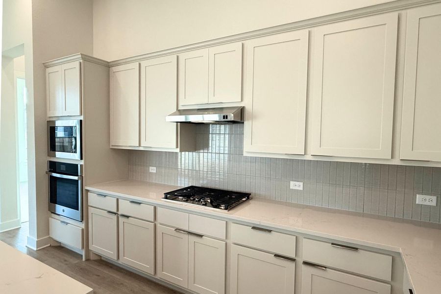 Contemporary kitchen featuring white shaker cabinetry, an integrated gas cooktop, double wall ovens, a coordinating stainless steel range hood, and light-toned countertops