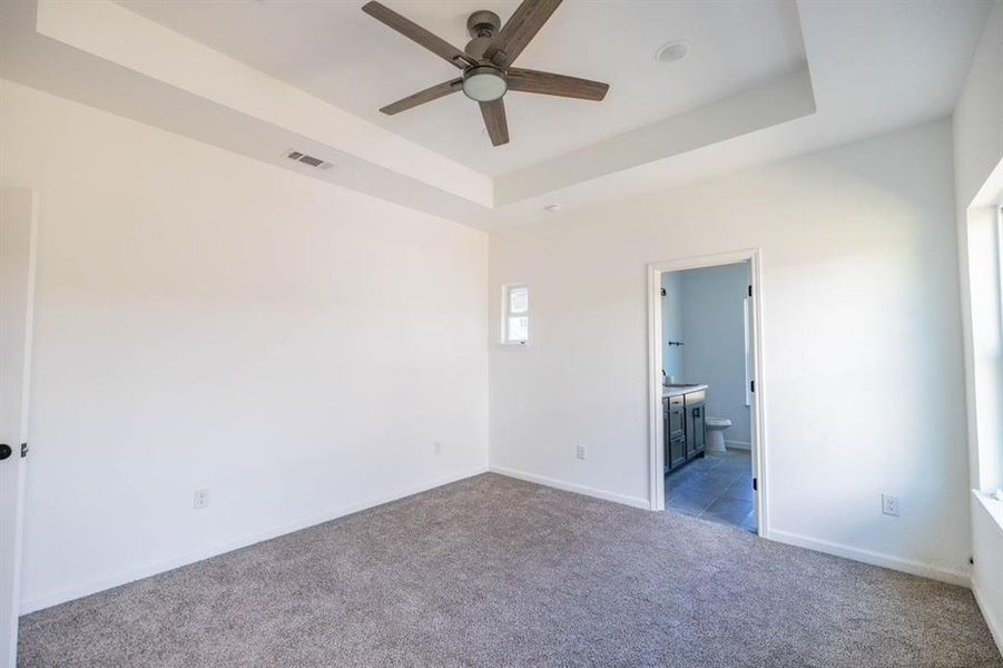 Unfurnished bedroom featuring ensuite bathroom, a tray ceiling, light colored carpet, and ceiling fan