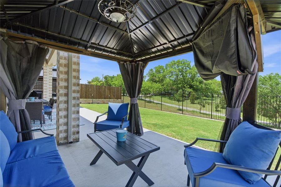Patio featuring a metal gazebo with an integrated ceiling fan, concrete flooring, and a manicured lawn beyond a black metal fence