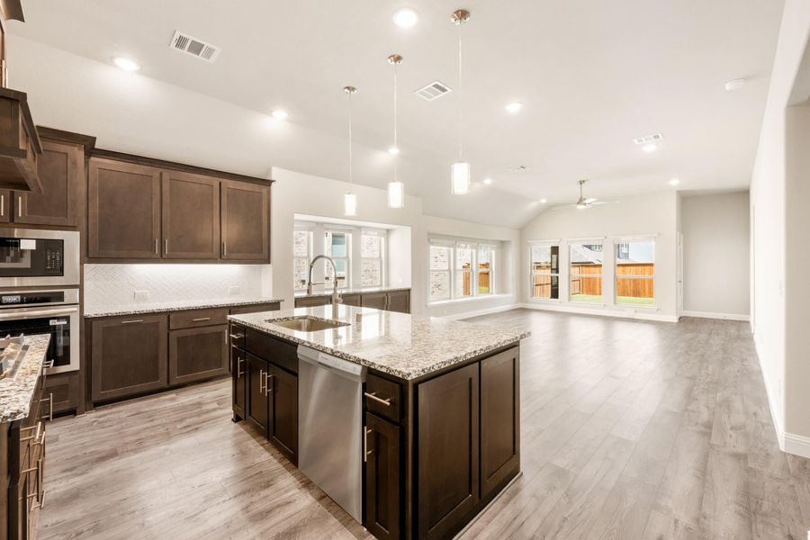 Open kitchen with dark brown cabinets, granite island, and light wood floors opening to living area