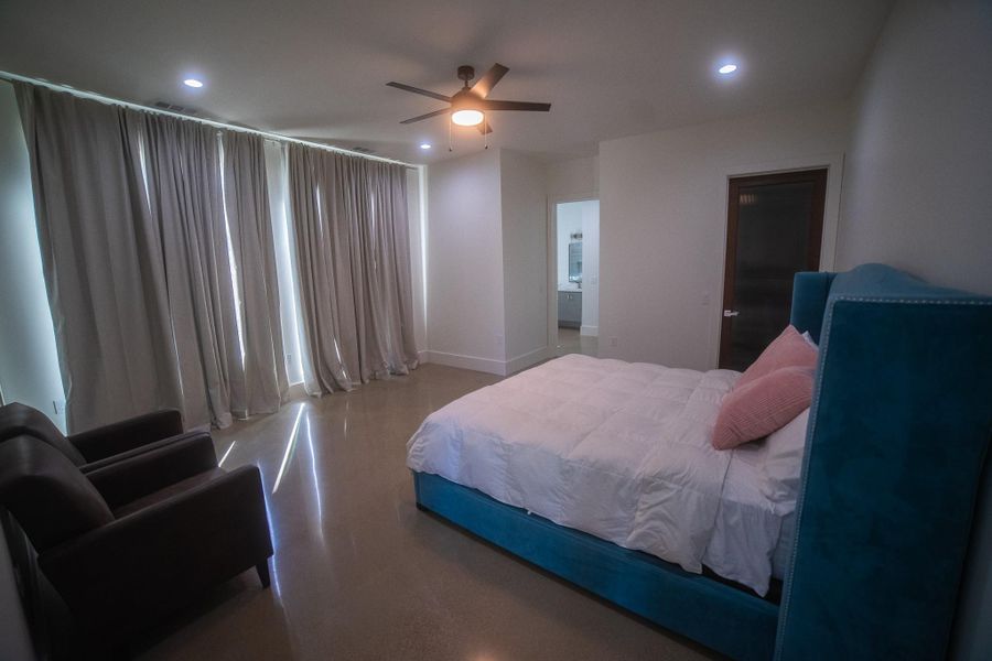 Spacious bedroom with polished floors, large windows with floor-length curtains, a ceiling fan, and a plush bed. It features two armchairs and an adjoining bathroom.