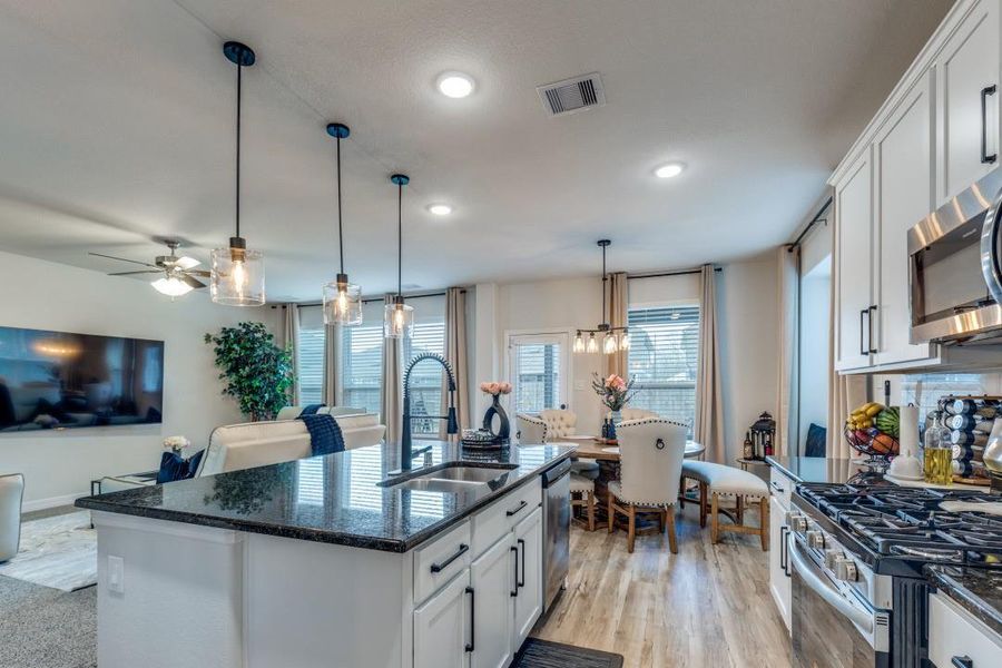 The space is bright and airy, featuring large windows and wood flooring, seamlessly flowing into a cozy living area with a ceiling fan and wall-mounted TV.