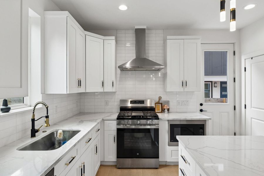 A close-up of the gourmet kitchen’s cooking wall. It features white shaker-style cabinetry with matte black hardware, a classic white subway tile backsplash, and high-end stainless steel GE appliances, including a gas range and a built-in microwave.