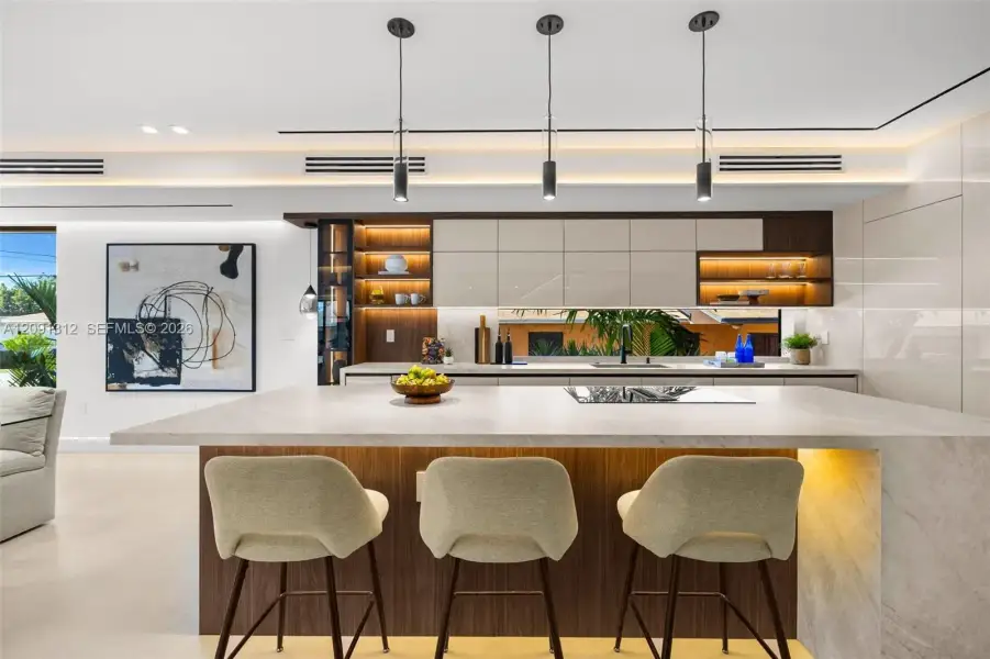 Kitchen featuring a large central island with stone-finish countertop, high-gloss cabinetry, linear pendant lighting, recessed accent shelving, and a horizontal backsplash window Kitchen featuring a large central island with stone-finish countertop, high-gloss cabinetry, linear pendant lighting, recessed accent shelving, and a horizontal backsplash window
