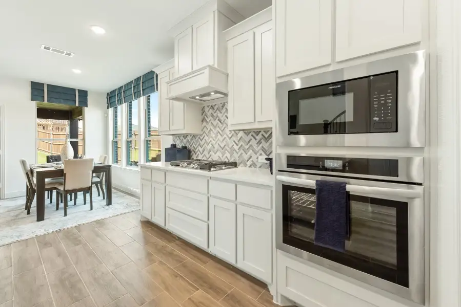 Kitchen with white cabinets, stainless steel built-in oven and microwave, herringbone tile backsplash, and dining area in background.