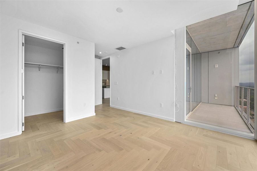 The spacious primary bedroom features elegant light wood herringbone flooring and clean, modern lines throughout. Floor-to-ceiling window allowing for abundant natural light. The room includes a generous walk-in closet with built-in shelving and hanging space. The spacious primary bedroom features elegant light wood herringbone flooring and clean, modern lines throughout. Floor-to-ceiling window allowing for abundant natural light. The room includes a generous walk-in closet with built-in shelving and hanging space.