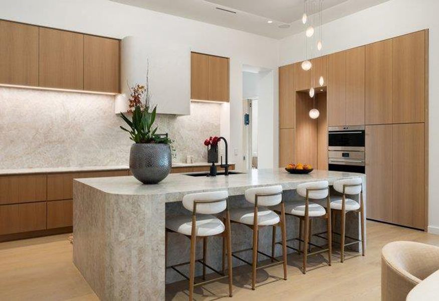 Gourmet kitchen featuring a waterfall island, integrated stainless steel appliances, light wood-finish cabinetry, a contemporary pendant light fixture, and a stone-finish backsplash
