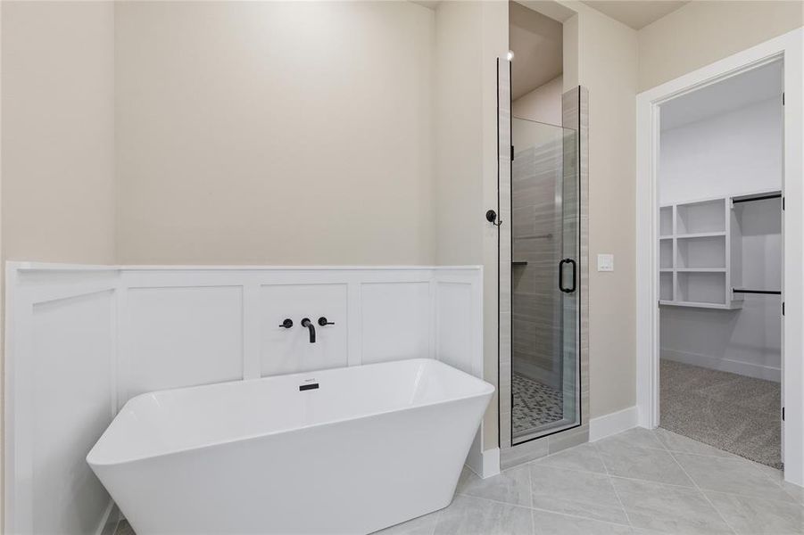 Freestanding soaking tub with wall-mounted matte black fixtures, glass-enclosed shower with tiled floor, and an adjacent walk-in closet featuring built-in shelving Freestanding soaking tub with wall-mounted matte black fixtures, glass-enclosed shower with tiled floor, and an adjacent walk-in closet featuring built-in shelving