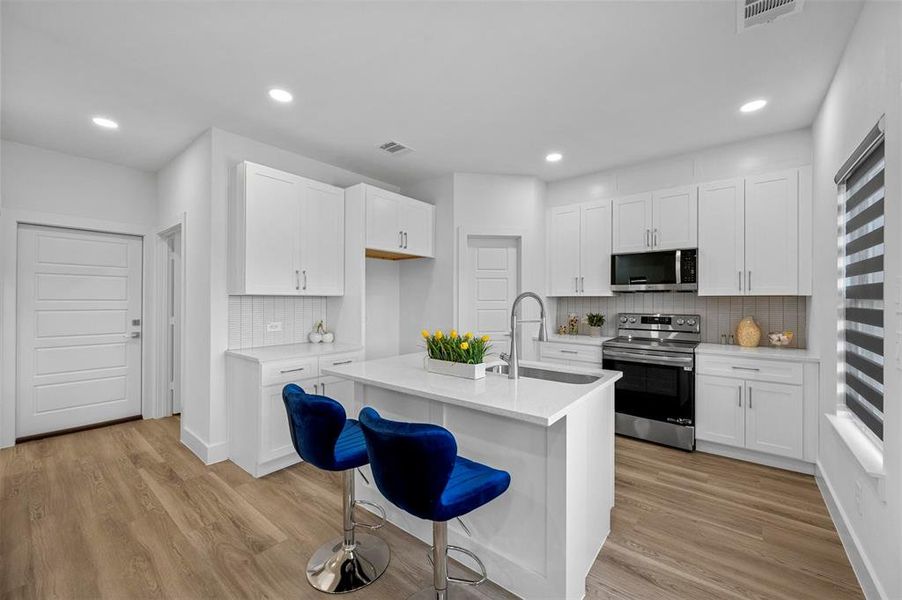 Kitchen with stainless steel appliances, a sink, white cabinets, light countertops, and a center island with sink