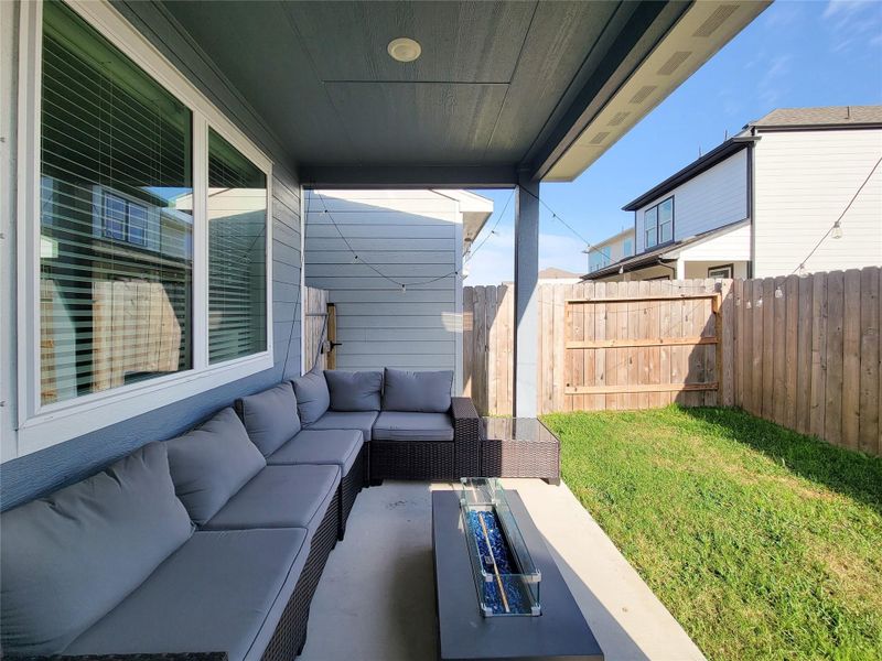 Cozy covered patio with a sectional sofa and a sleek fire pit table, perfect for outdoor relaxation. The fenced backyard offers privacy, with a small grassy area enclosed by wooden fencing.