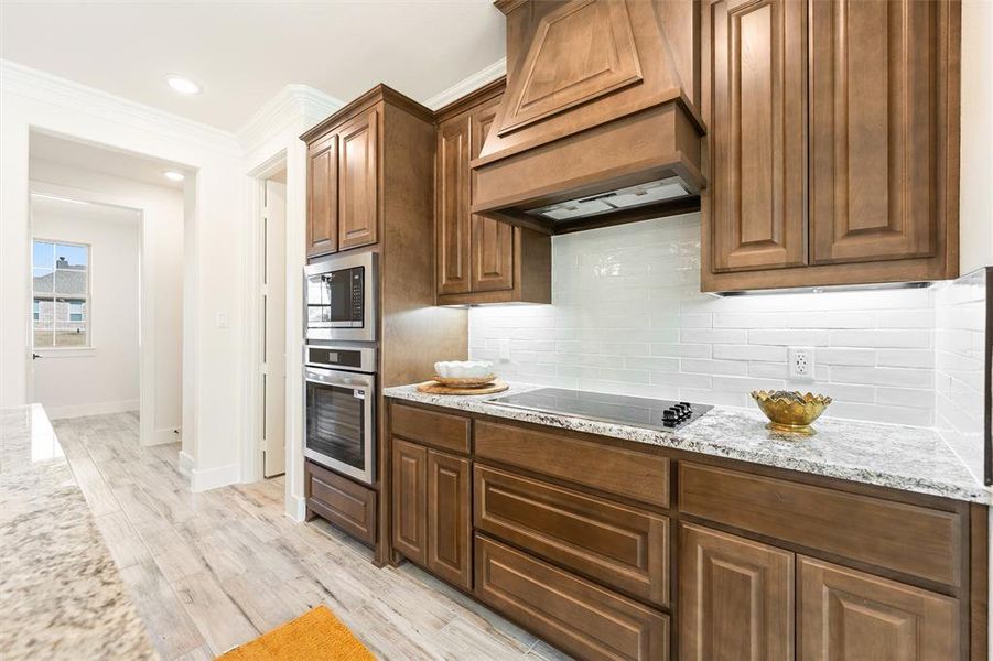 Kitchen featuring custom exhaust hood, light stone counters, appliances with stainless steel finishes, crown molding, and light wood-style floors