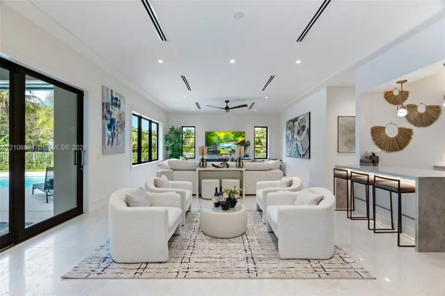 Open-concept living area featuring large-format stone flooring, recessed linear lighting, black-framed windows, integrated wet bar with stone countertop, and multi-panel sliding glass doors Open-concept living area featuring large-format stone flooring, recessed linear lighting, black-framed windows, integrated wet bar with stone countertop, and multi-panel sliding glass doors