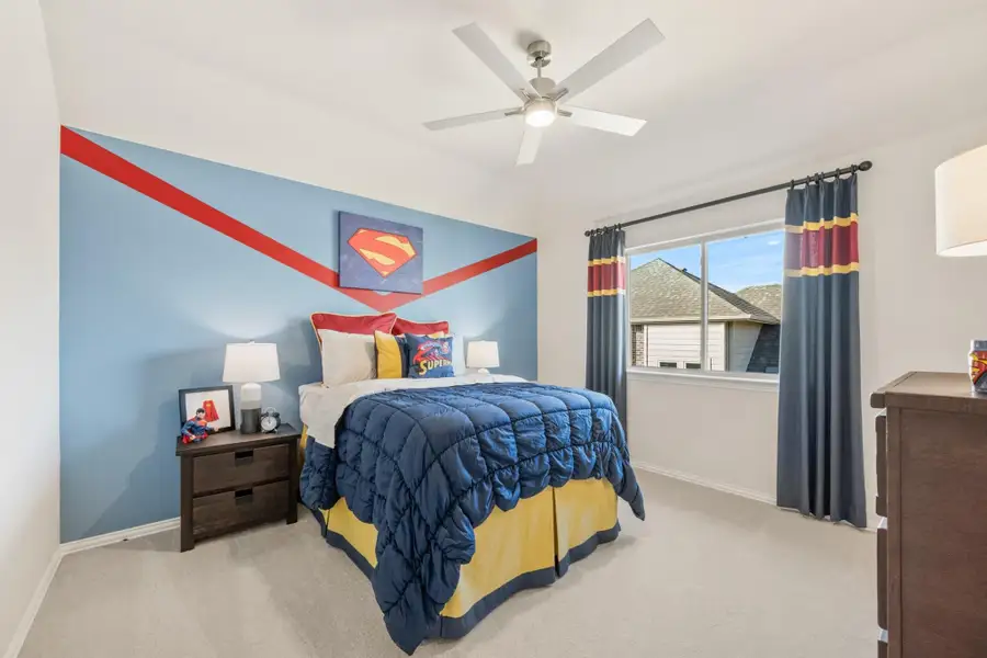Children's bedroom with Superman-themed accent wall, blue and yellow bedding, ceiling fan, and carpet flooring Children's bedroom with Superman-themed accent wall, blue and yellow bedding, ceiling fan, and carpet flooring