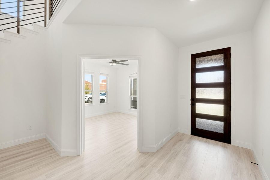 Bright entryway with dark wood glass-panel front door, light hardwood floors, and open view to adjacent room with ceiling fan.