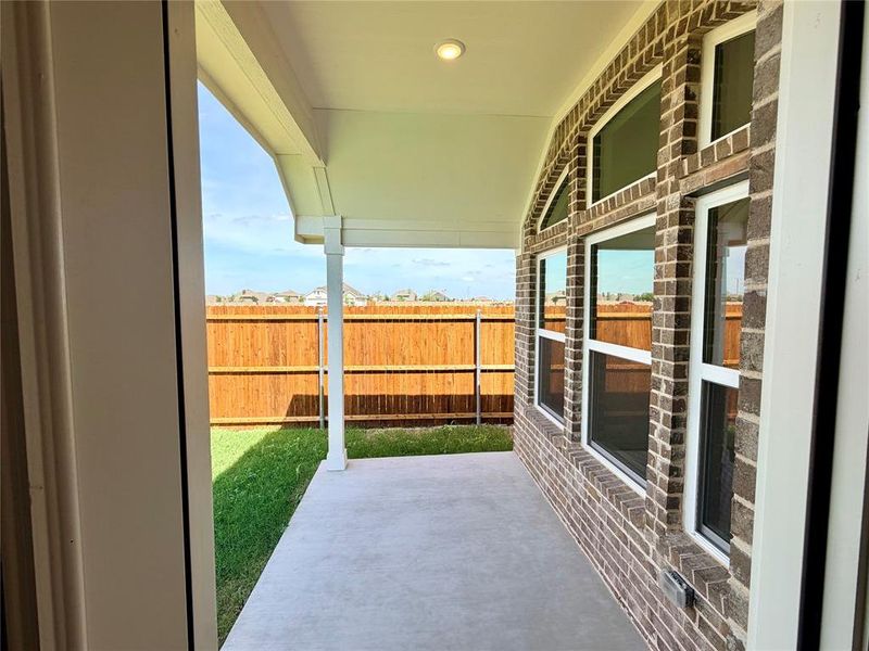 Covered patio area with a concrete surface, featuring a brick exterior wall with multiple windows, a recessed ceiling light, and a wooden privacy fence bordering a grass lawn