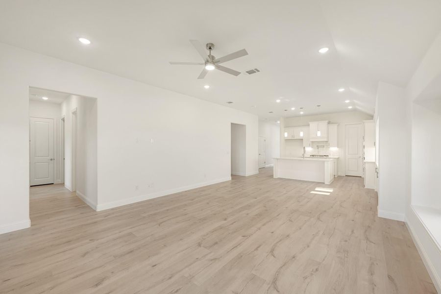 Open living area with light wood floors, ceiling fan, and white kitchen with island in background