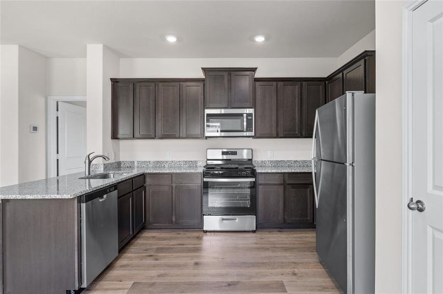 Kitchen featuring appliances with stainless steel finishes, light wood-style floors, light stone countertops, dark brown cabinetry, and a peninsula