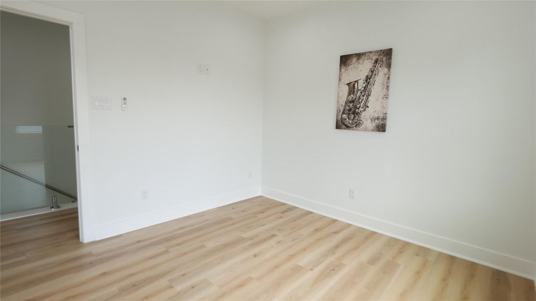 This room features light wood flooring and white walls, offering a bright and clean space. A piece of wall art adds a touch of character. The room has a modern feel with a visible glass railing and a doorway leading to an adjacent space. This room features light wood flooring and white walls, offering a bright and clean space. A piece of wall art adds a touch of character. The room has a modern feel with a visible glass railing and a doorway leading to an adjacent space.