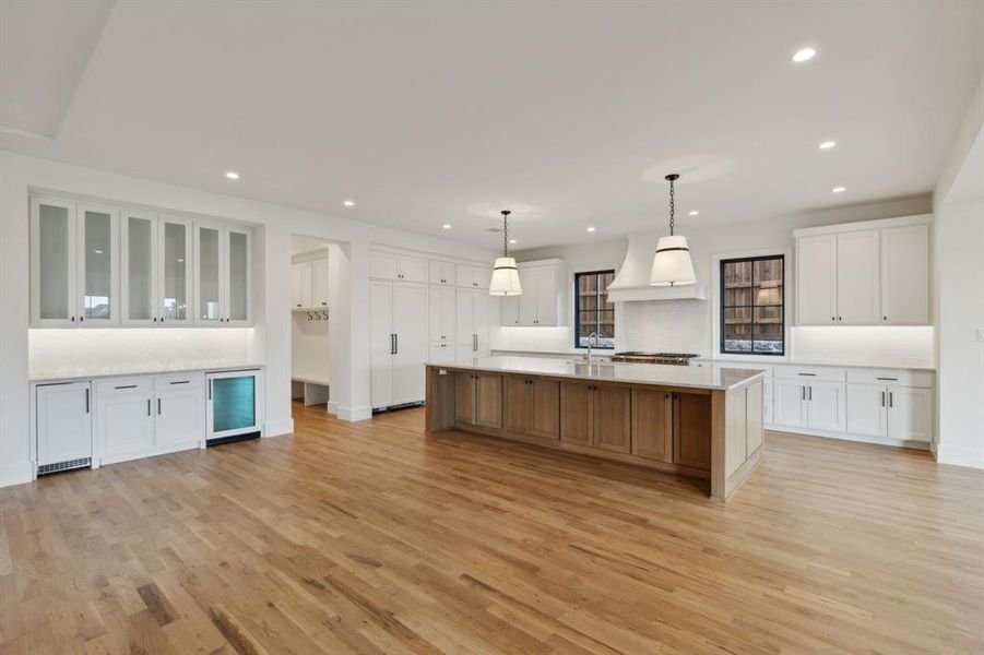 Kitchen featuring a large island with sink, white cabinets, hanging light fixtures, custom range hood, and light hardwood / wood-style flooring