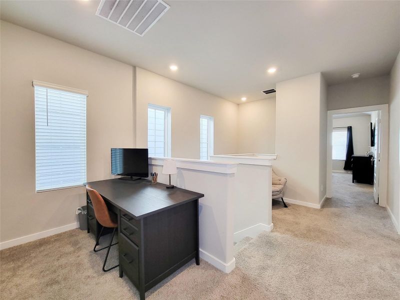 This photo shows a bright, carpeted home office space with a desk and chair, adjacent to a hallway leading to another room. It features recessed lighting and two windows with blinds for natural light.