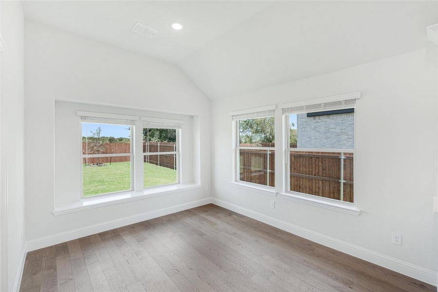 Empty room with vaulted ceiling, hardwood floors, and large windows overlooking a fenced backyard