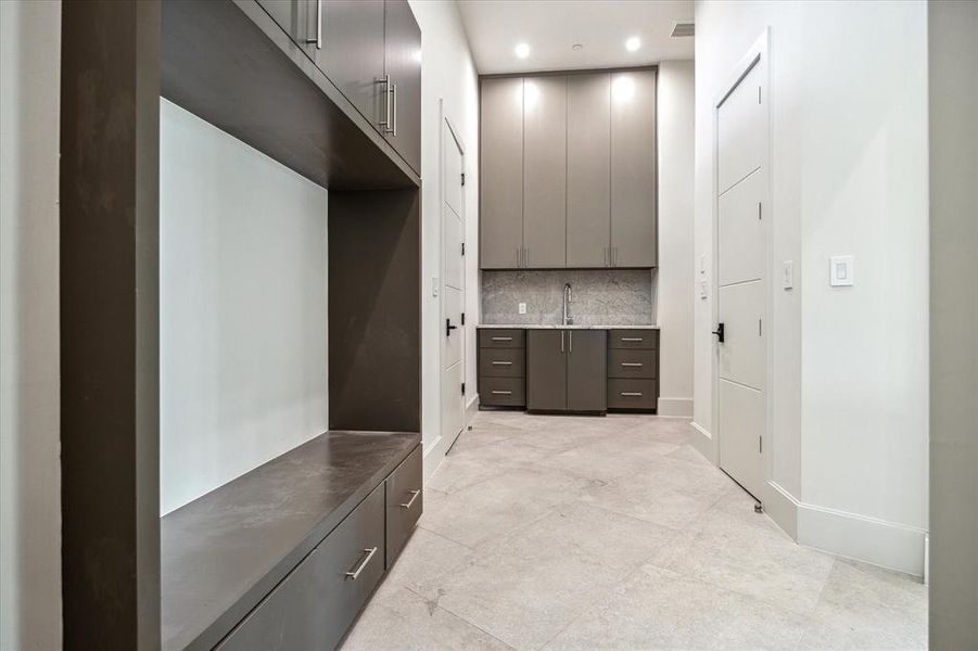 This luxurious mudroom features door access to a private garage, a wall of StevensWood cabinetry for ample storage, and a niche with a bench seat. The designer neutral wall paint and dune carved natural floor tile are complimementary creating a welcoming and functional space. The first floor utility room with Himalaya White countertops and backsplash, an Elkay undermount single bowl sink with a PROFLO pull-out spray faucet, and ample storage with upper and lower cabinetry, making laundry a breeze.