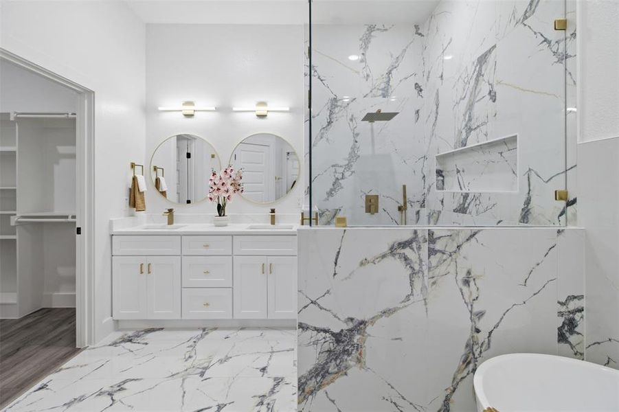 Spacious bathroom featuring marble-look flooring and shower surround, a dual vanity with white cabinetry and gold-tone hardware, two round mirrors with gold-tone frames, and a glass-enclosed shower with a rain shower head